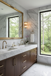Bathroom with marble countertop, wooden cabinets, and large window with view of trees. satin brass sconce with clear glass