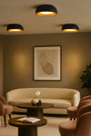 Modern living room with beige sofa, round wooden tables, and abstract art on the wall. Black and gold LED ceiling light.