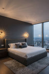 Modern bedroom with a large bed, dark walls, and city views. Glass wall sconce. 