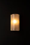 A frosted glass antique wall sconce with a satin brass backplate, illuminated, against a dark background.