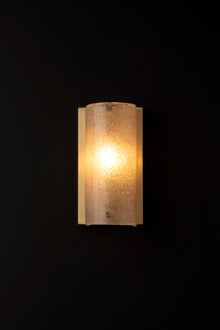  A frosted glass antique wall sconce with a satin brass backplate, illuminated, against a dark background.