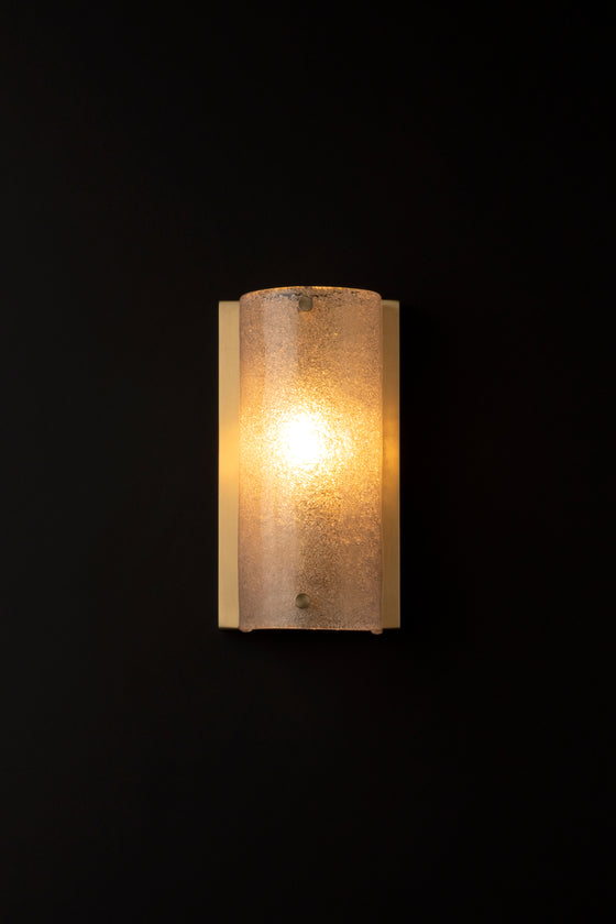 A frosted glass antique wall sconce with a satin brass backplate, illuminated, against a dark background.