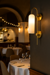Wall-mounted satin brass and opal glass lamp in a restaurant setting with tables and chairs.