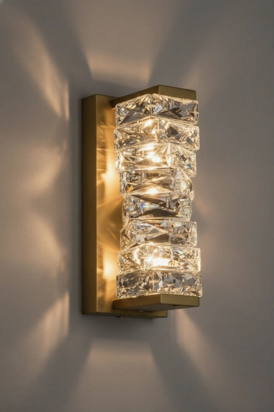 Wall-mounted satin brass LED light fixture with crystal-like glass panels on a gray wall.