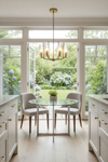 Modern kitchen with a round glass dining table and chairs, featuring a chandelier and large windows overlooking a garden.