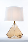 An amber geometric glass table lamp with a white fabric shade, featuring a satin brass base and a clear cord.