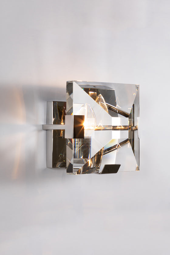 A contemporary polished stainless steel wall sconce with a clear glass lens and metallic frame, illuminated to showcase the lighting effect.