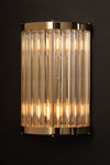 A wall-mounted sconce with a satin brass finish and clear glass rods, glowing warm white light.