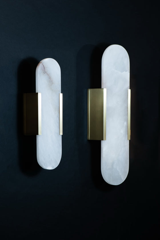 An oval-shaped wall sconce with a white alabaster shade and a satin brass finish, displaying a warm light LED glow.