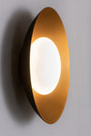 A modern wall light and ceiling fixture with a concave metal shade in black and satin brass finish, and a blown opal glass shade, emitting a warm white light.