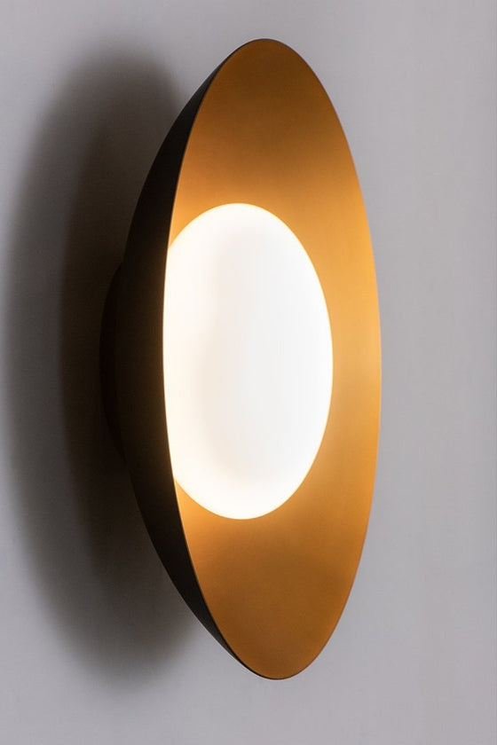 A modern wall light and ceiling fixture with a concave metal shade in black and satin brass finish, and a blown opal glass shade, emitting a warm white light.
