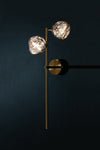 A wall sconce right facing with two globe-shaped lights, featuring a satin brass finish with black accents on the backplate, mounted on a dark wall.