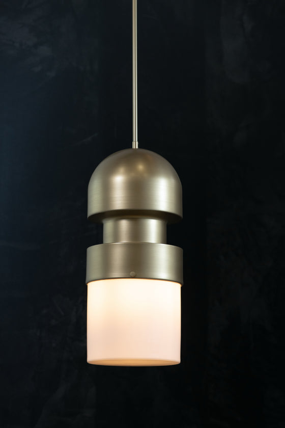 A modern pendant LED light with a satin brass finish and opal glass shade, hanging against a dark background.
