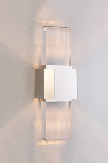  A polished nickel wall sconce with a simple geometric design featuring a beveled glass panel, emitting a bright array of light.