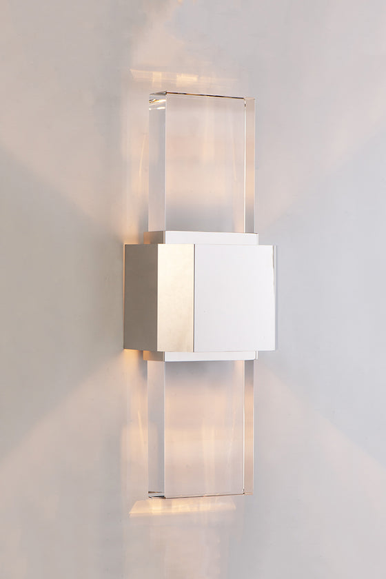 A polished nickel wall sconce with a simple geometric design featuring a beveled glass panel, emitting a bright array of light.